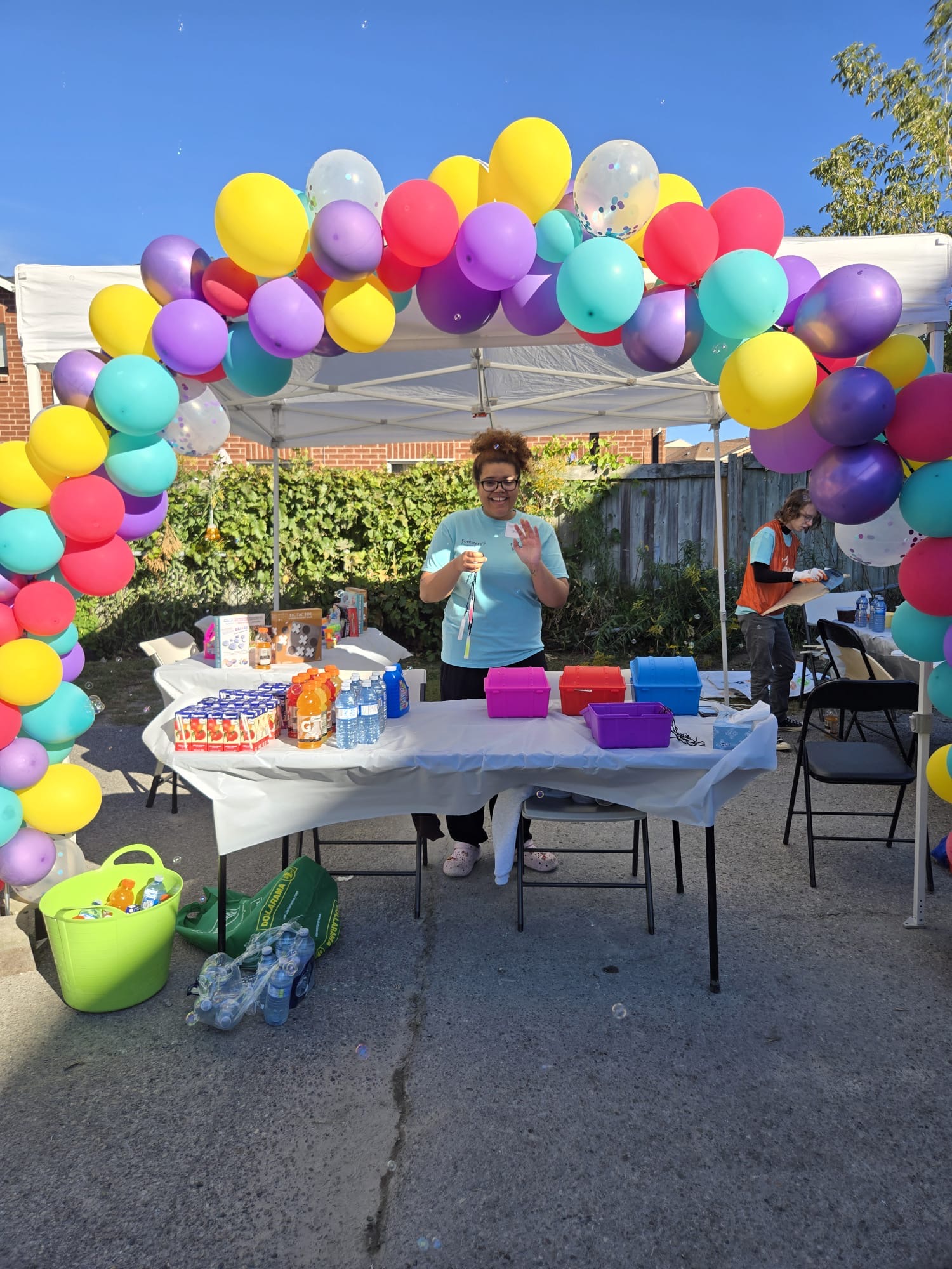 A volunteer blows bubbles behind a refreshments table under a white tent framed by a large arch of yellow, pink, purple and teal balloons at the Maple Glen playground build.