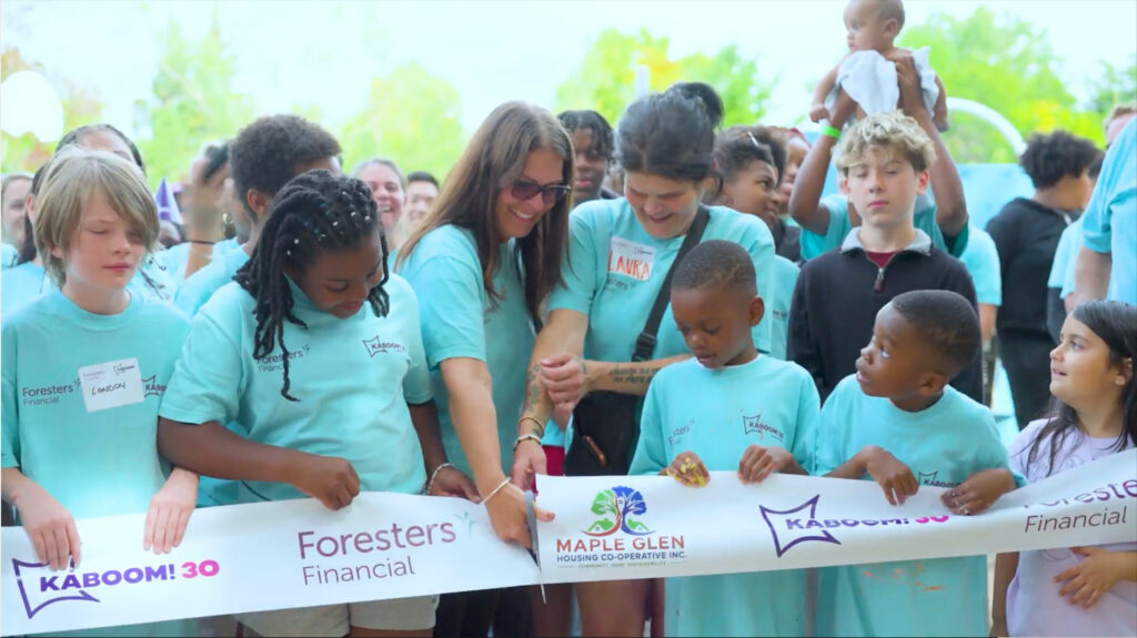 Children and adults in matching teal shirts hold a ribbon branded KABOOM! 30, Maple Glen Housing Co-operative and Foresters Financial while it is cut at the opening of the new playground in Oshawa.