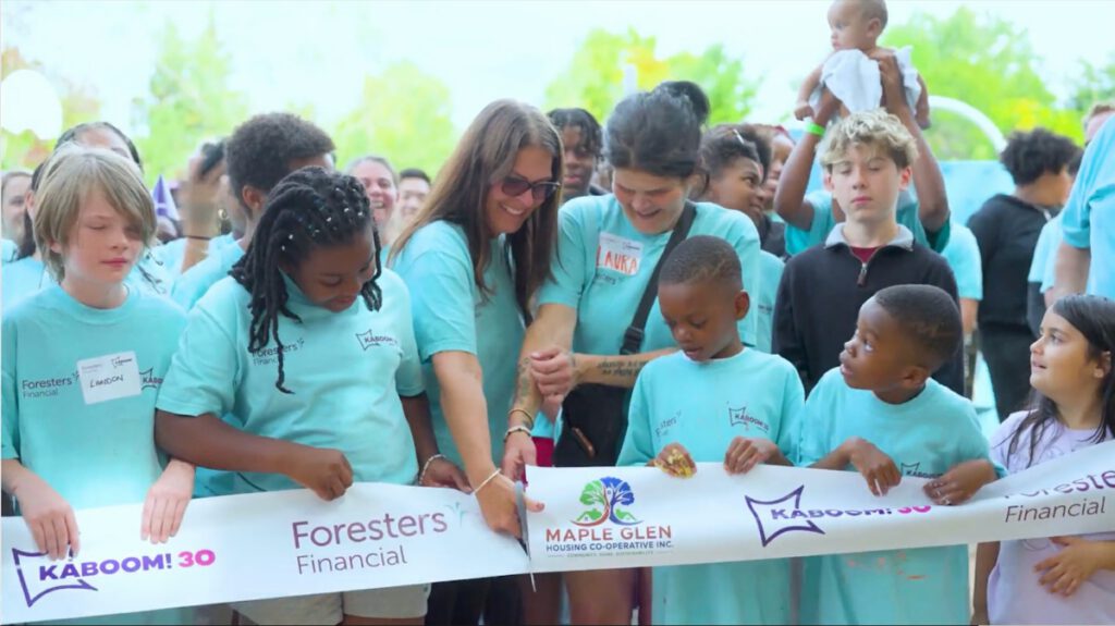 Children and adults in matching teal shirts hold a ribbon branded KABOOM! 30, Maple Glen Housing Co-operative and Foresters Financial while it is cut at the opening of the new playground in Oshawa.