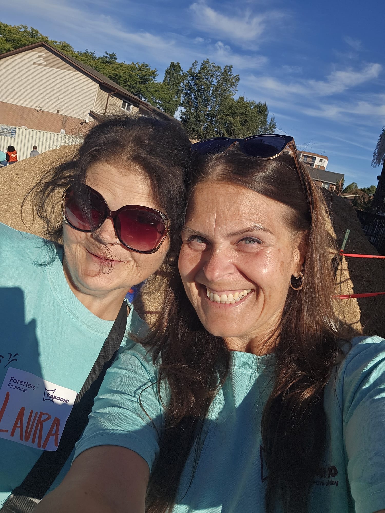 Two volunteers in teal Foresters Financial and KABOOM! shirts smile together for a photo in front of a large mound of wood chips destined for the playground safety surfacing.