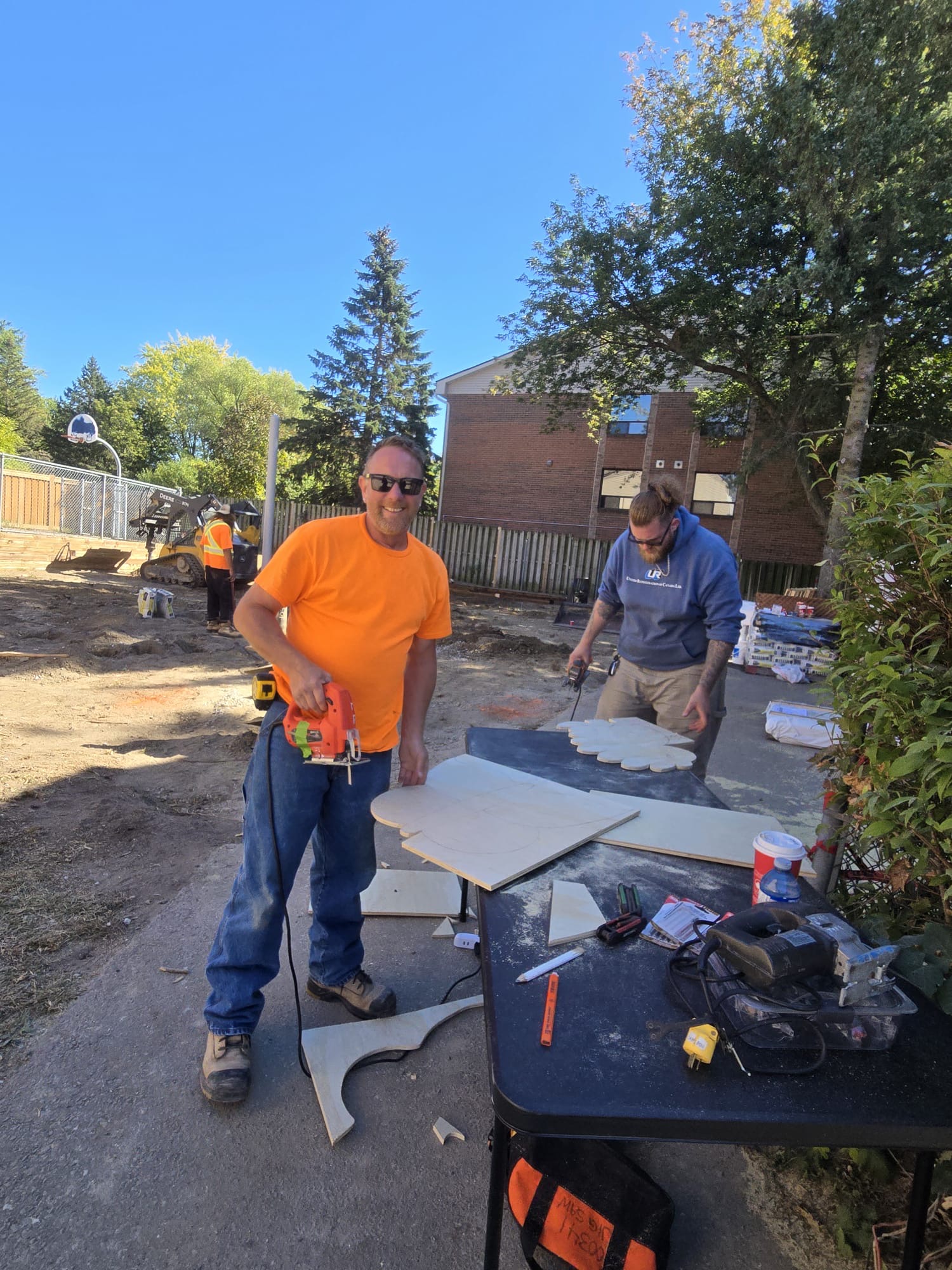 Two volunteers use jigsaws to cut curved plywood play panels on a folding table, with bare excavated ground and a skid steer behind them at the Maple Glen build site.