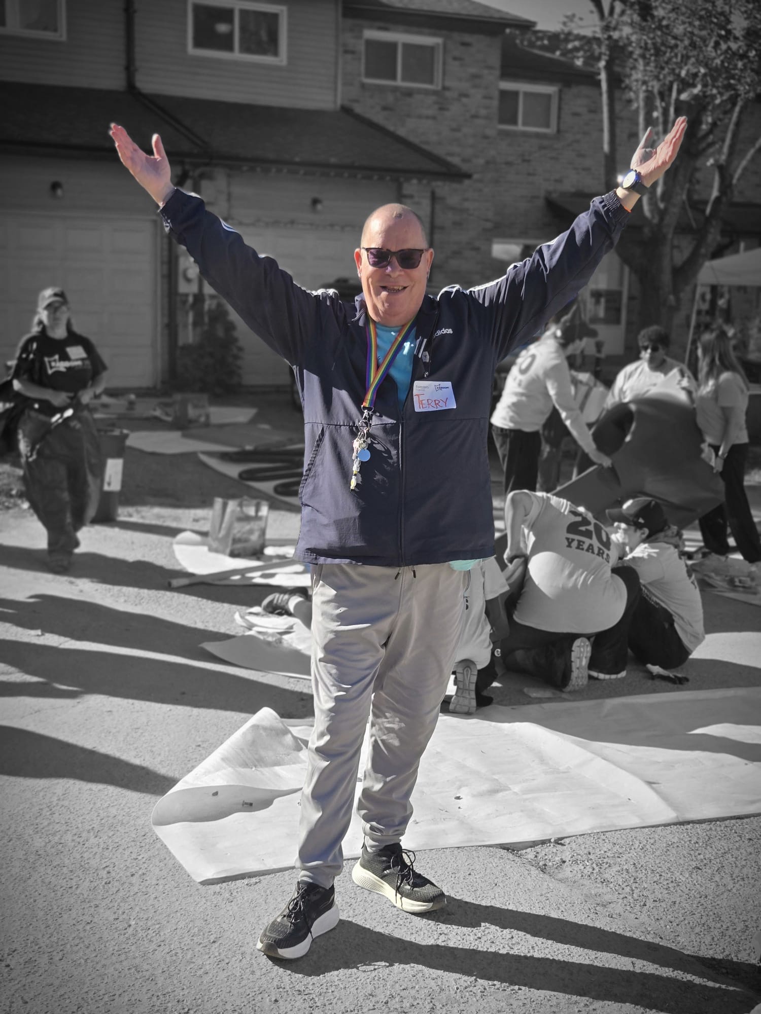 A volunteer with a Foresters Financial and KABOOM! name badge stands with both arms raised in celebration on build day, with other volunteers assembling play panels behind him.
